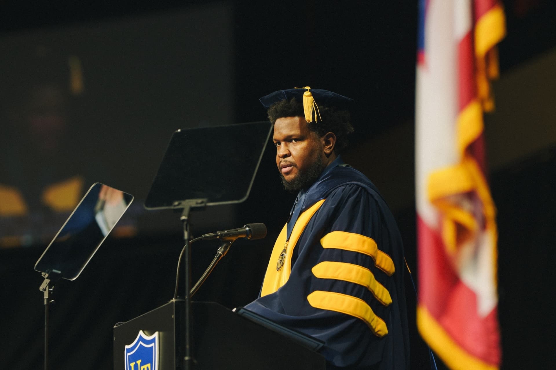 Will Lucas speaking at a university commencement in academic regalia.