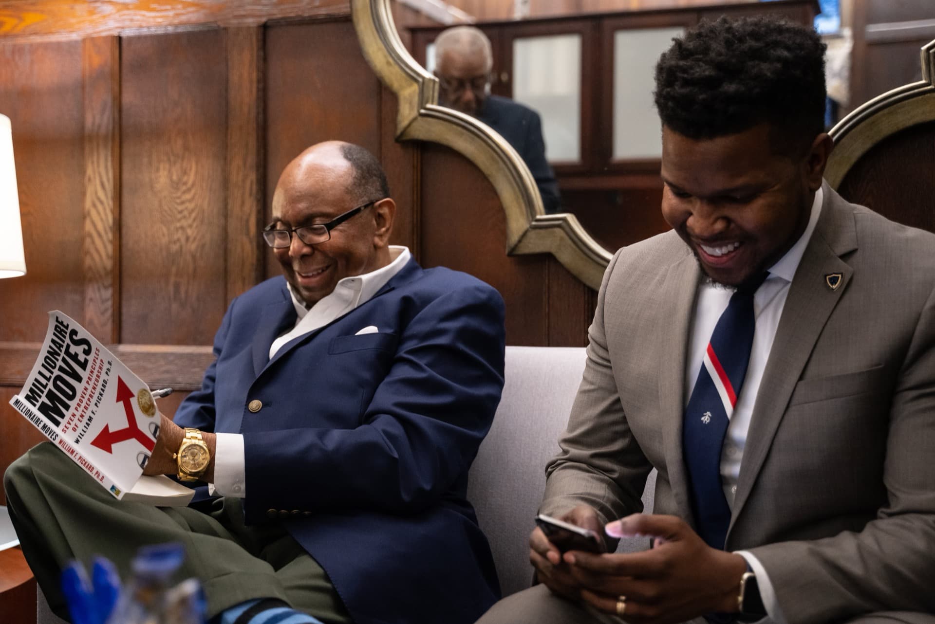 Will Lucas laughing on a couch beside Bill Pickard, who is holding a copy of his book Millionaire Moves.